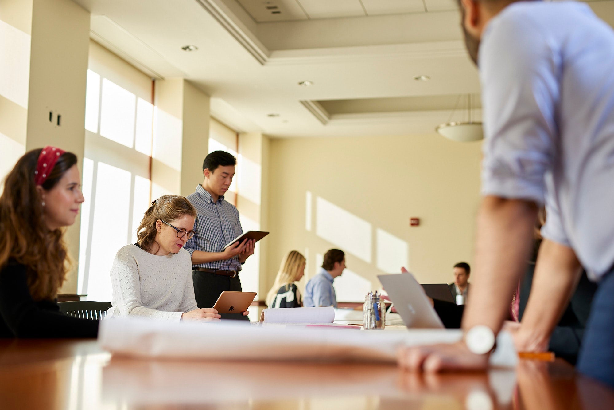 Steers students standing and sitting around a conference room table actively working on plans in front of them. Some are using laptops and tablets.