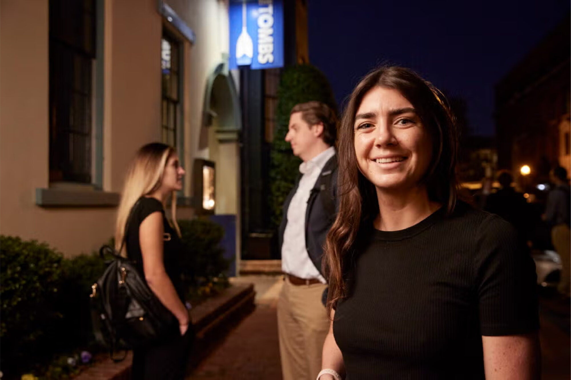 Three Steers alumni members outside in a evening gathering. One person in the forefront facing forward and smiling and two in the background speaking with one another.