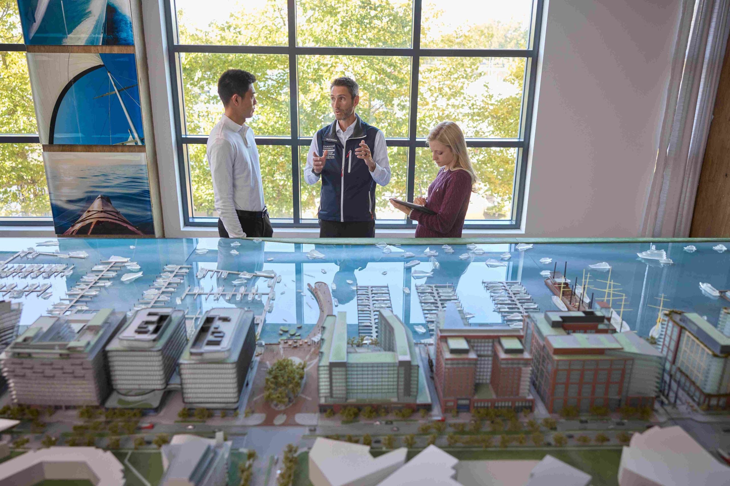 Director, Matthew Cypher and two Steers students stand around a detailed architectural model of a waterfront development. Large windows in the background let in natural light, illuminating the model and the group as they discuss.