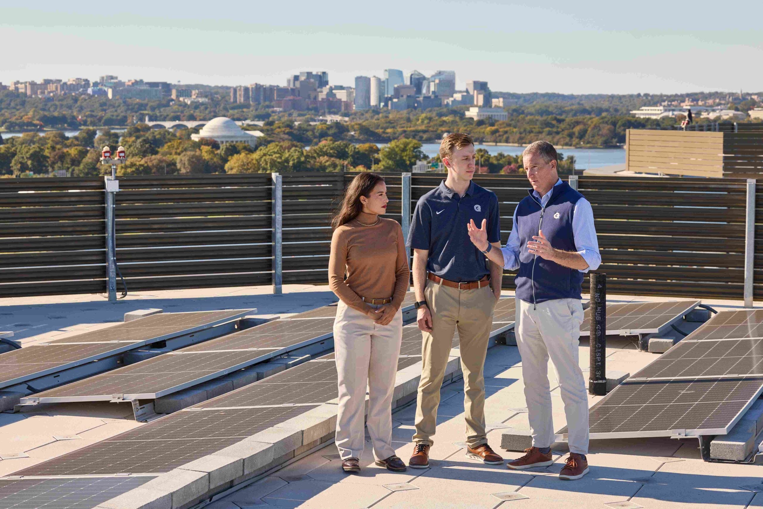 Three people talk outdoors on a rooftop, with a cityscape in the background.