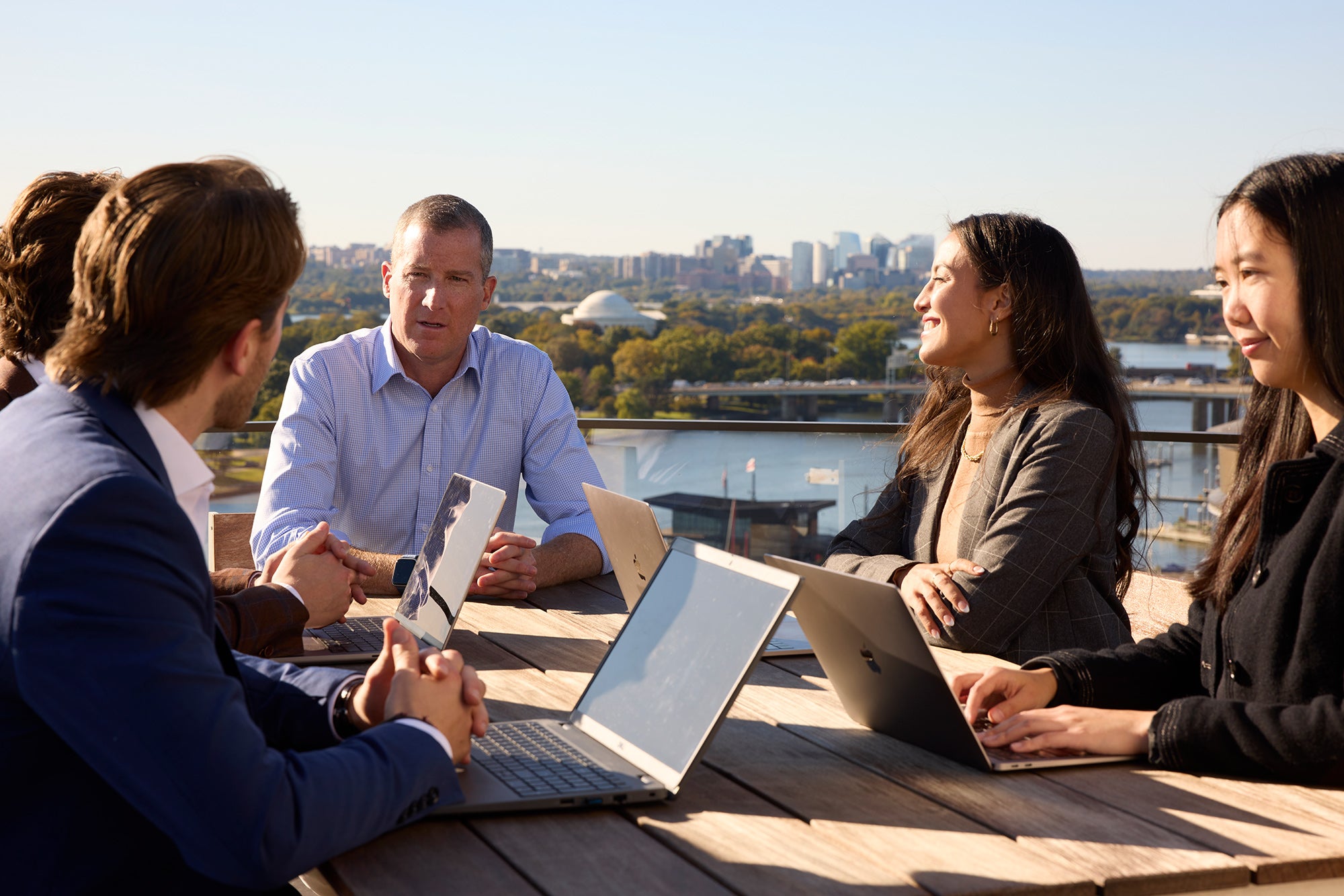 Executive Director, Tim Reardon sitting with four students at a table outside, chatting outside with a cityscape in the background.